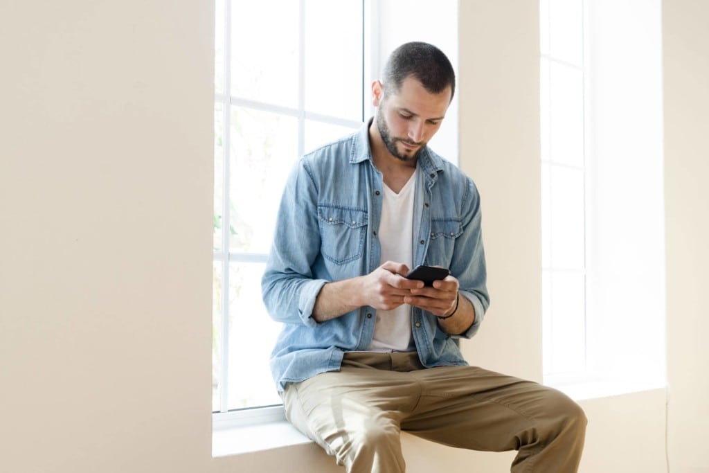Ein junger Mann schaut sitzt auf einem Fensterbrett und schaut ernst auf sein Smartphone
