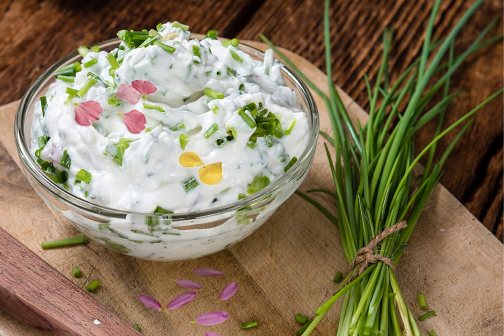 Ein Joghurt-Dipp mit Kräutern und Blüten in einer Schale. Daneben ein frisches Bund Schnittlauch.
